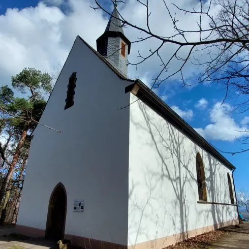 Eine kleine weiße Kirche steht umgeben von Bäumen unter einem blauen Himmel. Die Architektur zeigt einen schlichten und charmanten Stil.