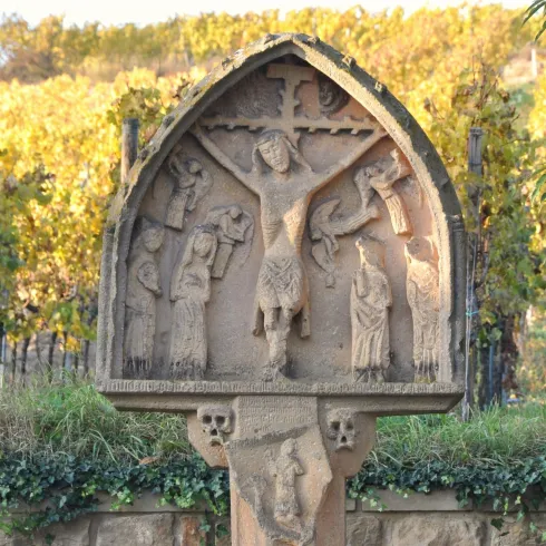 Eine kunstvolle Steinskulptur mit biblischen Motiven, die in einem Weinberg steht. Im Hintergrund sind grüne Weinreben und sanfte Hügel zu sehen.