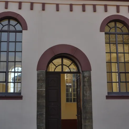 Eine historische Gebäudeansicht mit einer großen Holztür und hohen Fenstern. Die Fassade ist in sanften Farben gehalten, mit architektonischen Details über den Fenstern.