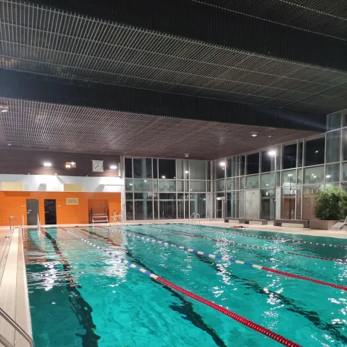 Ein gut beleuchtetes Schwimmbad mit klarem Wasser und roten Bahnmarkierungen. Die Umgebung ist modern mit großen Fenstern und Glaswänden.