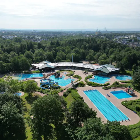 Ein gepflegtes Schwimmbadgebiet umgeben von Bäumen. Die Anlage verfügt über verschiedene Schwimmbecken und ist in einer grünen Umgebung eingebettet.