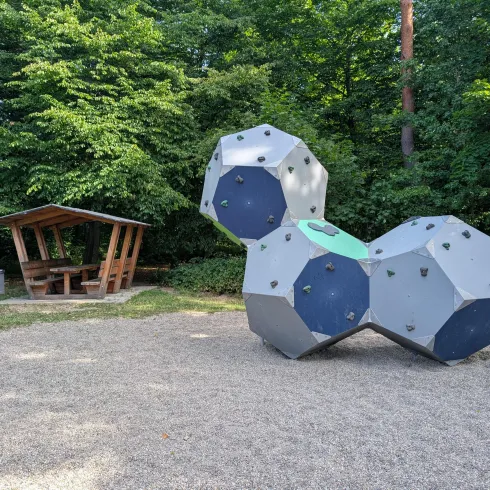 Ein Spielplatz mit einem kletterbaren Kletterelement und einer Überdachung aus Holz. Umgeben von dichtem Grün und Natur.