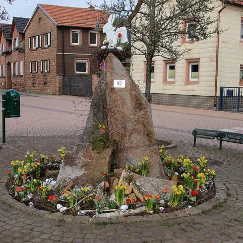 Ein hübscher Platz mit einem großen Stein in der Mitte, umgeben von bunten Blumen. Im Hintergrund sind Häuser und eine Straßenansicht sichtbar. Hier ist der Dorfbrunnen der Gemeinde Esthal zu sehen.