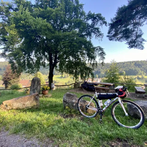 Ein Gravelbike steht an einem Sandstein angelehnt, vor einem schönen Ausblick auf eine grüne Landschaft. Im Hintergrund sind Bäume und Sitzgelegenheiten zu sehen.