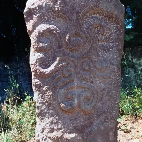 Ein großer, aufrechter Stein mit kunstvollen, spiralförmigen Mustern. Die Umgebung ist grün und natürlich, was die historische Bedeutung des Steins hervorhebt.
