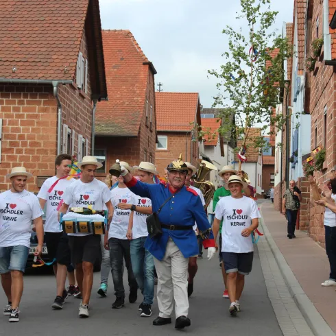 Eine Gruppe von Menschen in weißen T-Shirts marschiert durch eine malerische Straße. Im Hintergrund sind historische Häuser und eine Frau, die zuschaut.