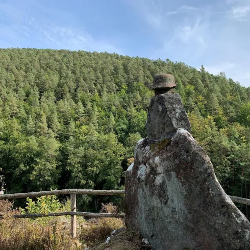 Ein großer Felsen mit einer charakteristischen Form steht vor einem bewaldeten Hang. Der Himmel ist klar und die Landschaft wirkt ruhig und friedlich.