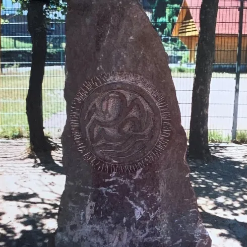 Ein großer, roter Stein mit einer dekorativen Gravur. Im Hintergrund sind Bäume und ein Gebäude sichtbar.