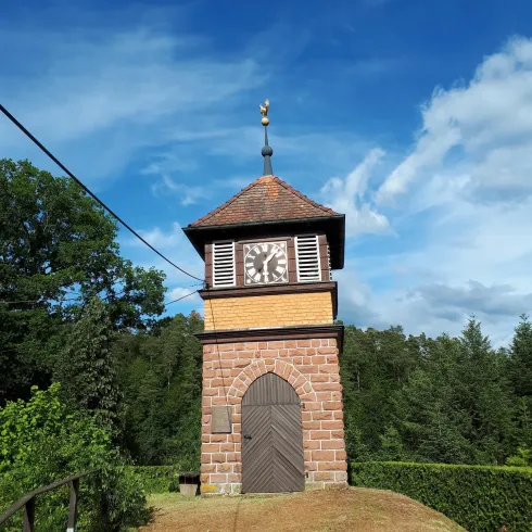 Ein schöner Glockenturm mit einem steilen Dach steht auf einer Wiese. Im Hintergrund sind grüne Bäume und ein blauer Himmel sichtbar.