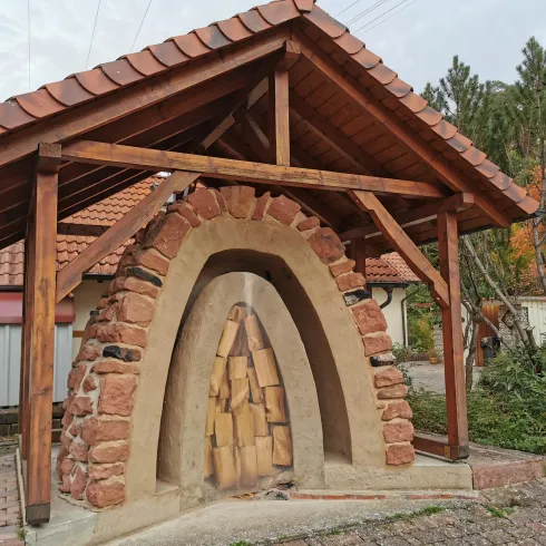Ein Holzunterstand mit einem gemauerten Kamin aus Steinen und Holz. Der Bau hat ein schützendes Dach und befindet sich in einer grünen Umgebung.