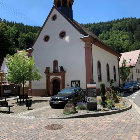 Eine kleine Kirche mit einem markanten Dach in einem ruhigen Dorf. Umgeben von Bäumen und gepflasterten Wegen.