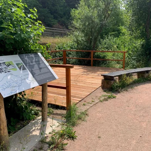 Ein ruhiger Aussichtspunkt mit einer Holzplattform und einer Bank. Umgeben von grüner Vegetation und einem Kiesweg.