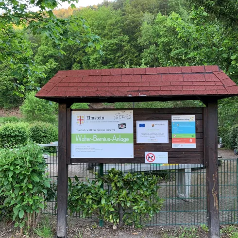 Ein Informationsstand im Grünen mit einem Dach. Daran sind verschiedene Schilder angebracht, die Besucher willkommen heißen. Die Infowand steht am Eingang zur Walter-Bernius-Anlage.