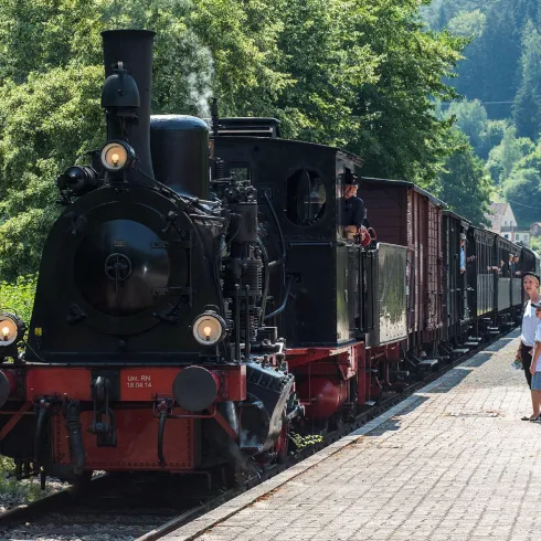 Eine alte Dampflok steht an einem Bahnhof. Passagiere warten auf die Abfahrt und genießen die Umgebung.