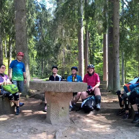 Eine Gruppe von Radfahrern sitzt um einen Tisch in einem Wald. Die Sonne scheint durch die Bäume und die Stimmung ist entspannt.