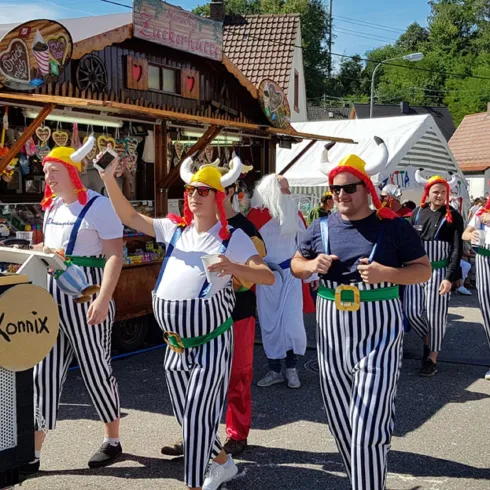 Eine feierliche Parade mit verkleideten Teilnehmern in gestreiften Hosen und bunten Hüten. Im Hintergrund sind verschiedene Buden und typisch deutsche Gebäude sichtbar.