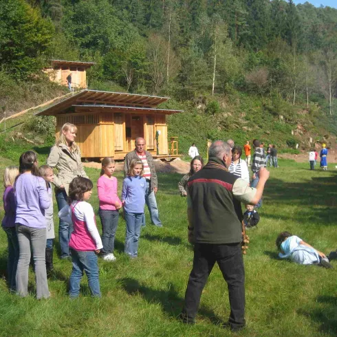 Eine Gruppe von Kindern und Erwachsenen steht auf einer Wiese. Im Hintergrund sind hölzerne Gebäude und Bäume zu sehen.