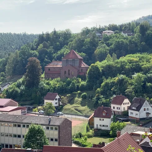 Eine malerische Landschaft mit einem Hügel, auf dem eine Kirche steht. Im Vordergrund sind mehrere Häuser zu sehen, umgeben von Grün.