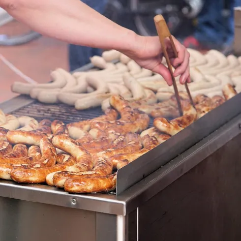 Eine Grillstation mit verschiedenen Würsten auf dem Grill. Eine Hand wendet die gegrillten Würste in der rauchigen Atmosphäre.