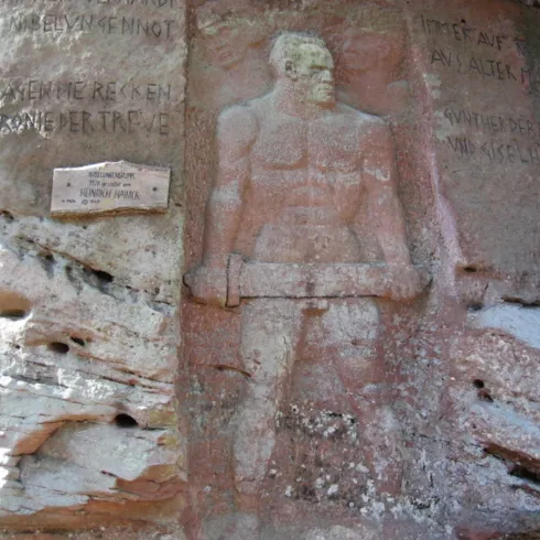 Eine Felswand mit einer kunstvollen Skulptur eines muskulösen Mannes, der einen Hammer hält. Über der Statue sind Inschriften zu sehen.