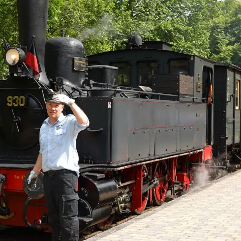 Die dampfbetriebene Lokomotive des Kuckucksbähnels steht am Kuckucksbahnhof in Elmstein. Ein Arbeiter begrüßt die Passagiere neben den Waggons in einer grünen Umgebung.
