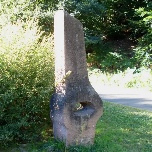 Ein auffälliger Stein mit einer durchbrochenen Form steht in der Natur. Er ist von Büschen umgeben und befindet sich in der Nähe einer Straße.