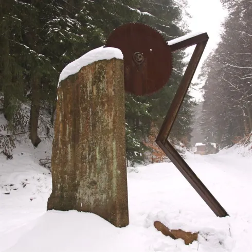 Eine Skulptur aus Stein und Metall steht im verschneiten Wald. Der Unterschied zwischen den Materialien und die winterliche Umgebung erzeugen eine interessante Atmosphäre.