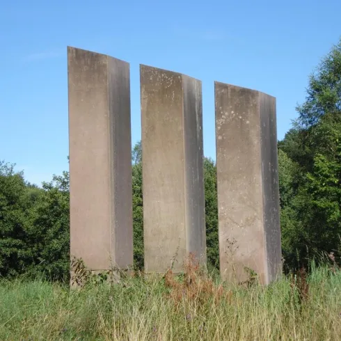 Drei hohe, rechteckige Steinsäulen stehen in einer grünen Wiese. Im Hintergrund sind Bäume und ein klarer blauer Himmel zu sehen.