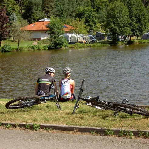 Zwei Radfahrer sitzen am Ufer eines ruhigen Sees und genießen die Aussicht. Ihre Fahrräder liegen neben ihnen auf dem Gras.