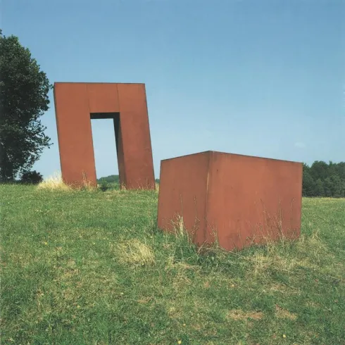 Eine moderne Skulptur aus rostigem Metall auf einer grünen Wiese. Im Hintergrund ist ein Baum zu sehen und der Himmel ist klar.