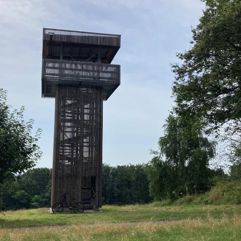 Ein Aussichtsturm steht auf einer Wiese, umgeben von Bäumen. Der Himmel ist teilweise bewölkt und es wirkt ruhig und friedlich.