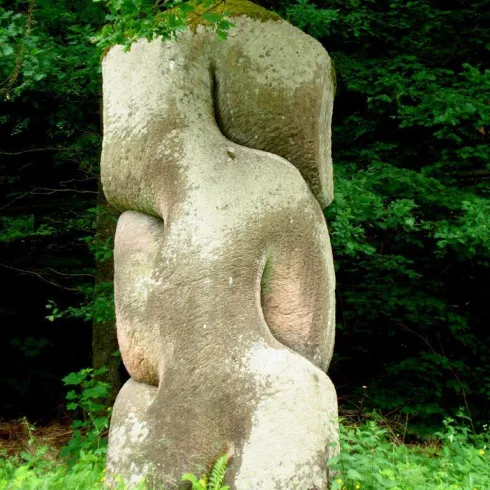 Eine stilisierte Steinskulptur steht inmitten von üppigem Grün. Die Form des Steins erinnert an menschliche Silhouetten.
