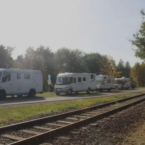 Ein Wohnmobilparkplatz neben einer Bahnlinie. Im Hintergrund sind Bäume und grünes Gras zu sehen.