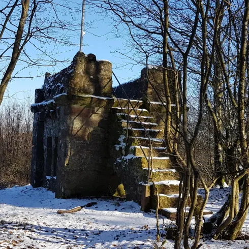 Eine verlassene Ruine mit einer Treppe, umgeben von kahlen Bäumen. Der Boden ist mit Schnee bedeckt und der Himmel ist klar.
