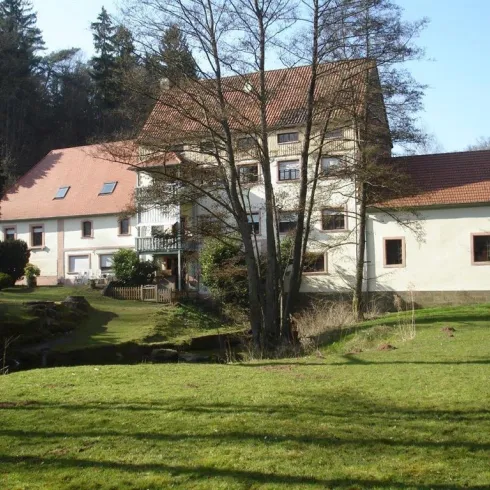 Ein großes Gebäude mit rotem Dach umgeben von Bäumen und einer grünen Wiese. Das Haus hat mehrere Fenster und einen ruhigen Gartenbereich.