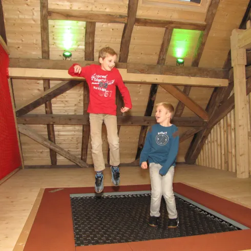 Zwei Kinder springen auf einem Trampolin in einem Holzhäuser. Der Raum ist gemütlich und gut beleuchtet.