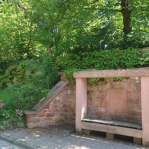 Eine ruhige Sitzbank aus Stein in einer grünen Umgebung. Bäume und Sträucher umgeben den Bereich und schaffen eine einladende Atmosphäre.