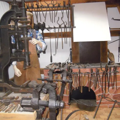 Eine Werkstatt mit traditionellen Schmiedewerkzeugen und einem großen Amboss. Im Hintergrund ist ein Schmied zu sehen, der an einem Projekt arbeitet.