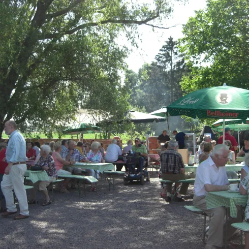 Eine lebhafte Gartenrestaurant-Szenerie mit vielen Gästen. Tische unter Bäumen und Sonnenschirmen schaffen eine einladende Atmosphäre.