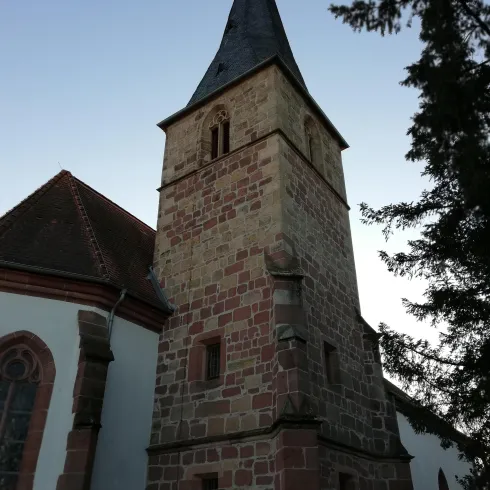 Ein historischer Kirchturm aus Stein mit einem spitzen Dach. Umgeben von Bäumen und einem klaren Himmel.