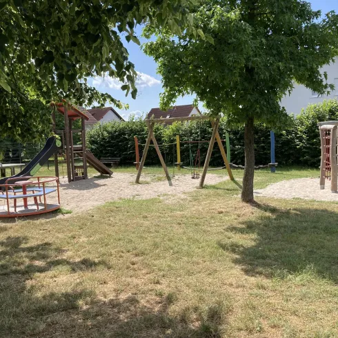 Ein Spielplatz mit Rutsche, Schaukeln und Klettergerüst. Der Bereich ist von Bäumen umgeben und bietet viel Grasfläche.