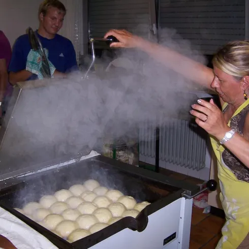 Eine Frau in einer Küche sprüht Wasser auf dampfende Teigbälle. Im Hintergrund steht ein Mann, der zuschaut.