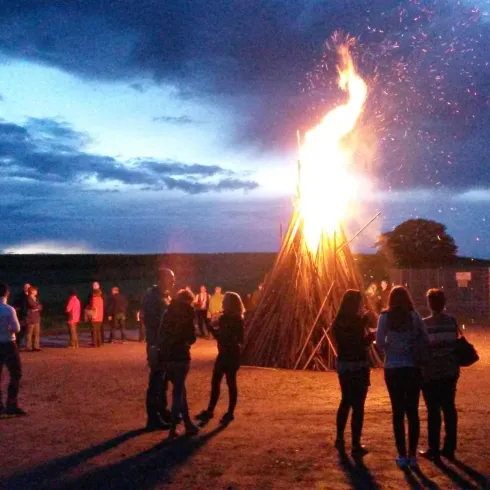 Ein großes Lagerfeuer brennt in der Dämmerung, umgeben von einer Gruppe von Menschen. Der Himmel ist bewölkt, und die funkelnden Glutpartikel fliegen in die Luft.