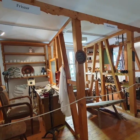 Ein historischer Friseursalon mit Holzwänden und alten Möbeln. Regale mit verschiedenen Gegenständen und Frisurausstattung sind sichtbar.