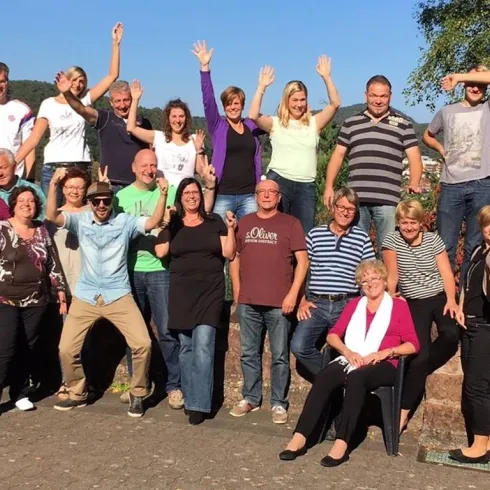 Eine Gruppe von Menschen steht fröhlich draußen in der Sonne. Sie posieren in verschiedenen Stellungen und zeigen ihre Begeisterung.