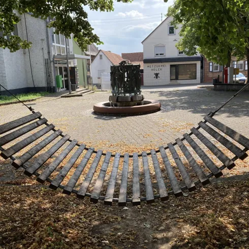 Eine hängende Holzschaukel in einem ruhigen Platz mit Bäumen. Im Hintergrund steht ein Brunnen und einige Gebäude sind sichtbar.