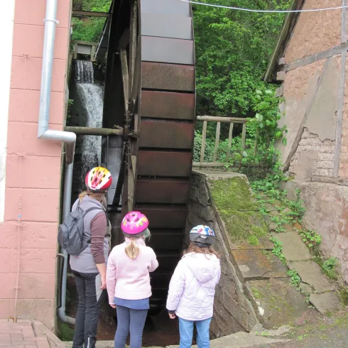 Ein Wasserrad, das an einem kleinen Bach steht, mit drei Personen in Schutzhelmen, die es beobachten. Im Hintergrund sind Bäume und eine alte Mauer zu sehen.