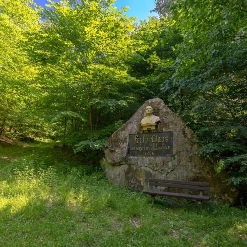 Ein Denkmal aus Stein steht in einem grünen Wald. Umgeben von Bäumen und Wiese vermittelt die Szene eine ruhige Naturatmosphäre.