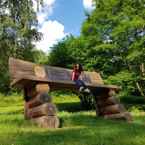 Eine Frau sitzt auf einer großen Holzbank im Freien. Im Hintergrund sind grüne Bäume und ein blauer Himmel zu sehen.