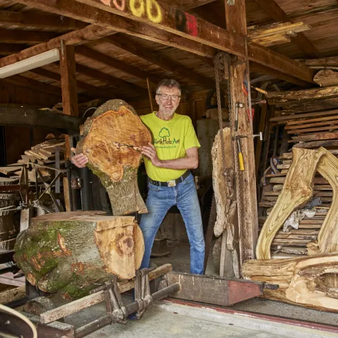 Ein Mann steht in einer Werkstatt mit einem großen Holzstück. Im Hintergrund sind weitere Holzstücke und Werkzeuge sichtbar.
